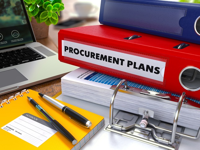 Binders and notebooks on a crowded desk. One of the binders’s spine reads Procurement Plans.